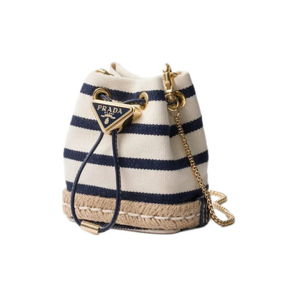 Prada canvas bucket bag with navy and cream horizontal stripes, a beige jute-wrapped base, a gold-tone triangular Prada logo charm, and a gold chain strap on a white background.