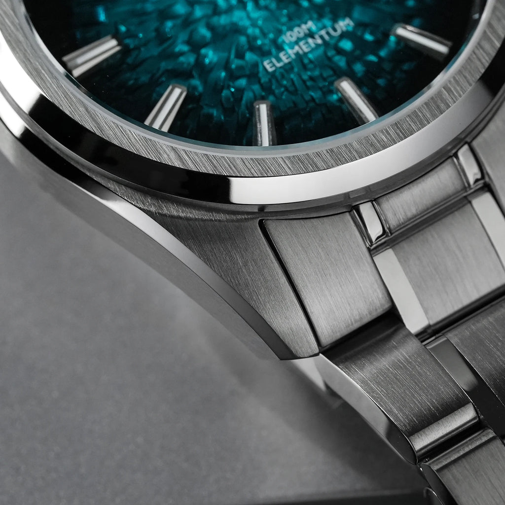 Close-up of a stainless steel watch with a textured teal-blue dial and matching metal bracelet, showing the brand text 'ELEMENTUM' on the face.