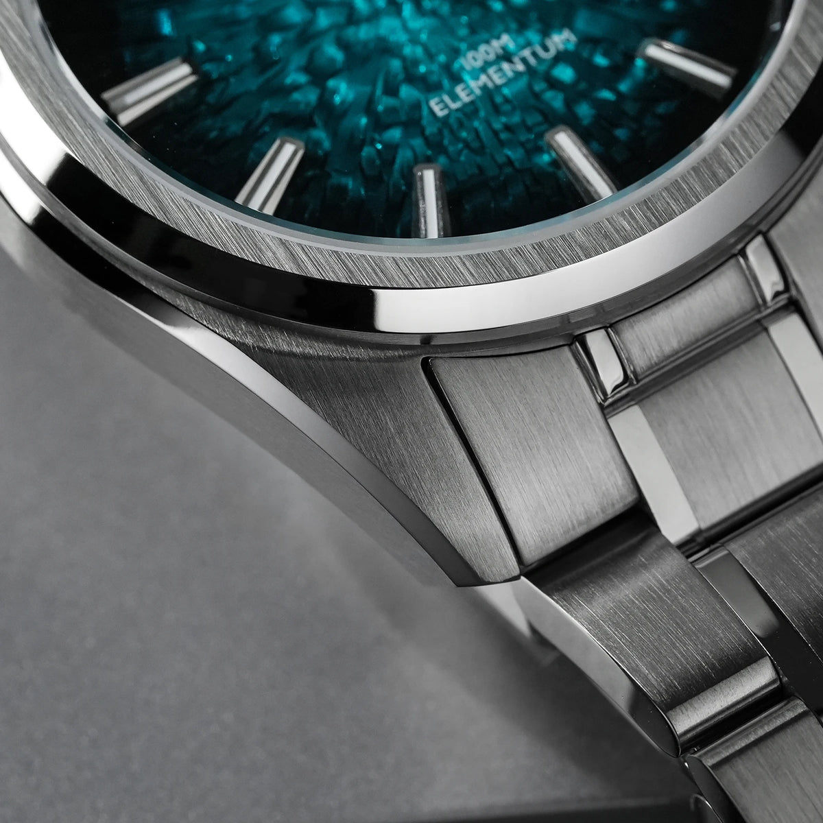 Close-up of a stainless steel watch with a textured teal-blue dial and matching metal bracelet, showing the brand text 'ELEMENTUM' on the face.