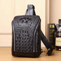 Black crocodile-embossed leather crossbody bag with a front zip pocket and shoulder strap, displayed on a wooden surface next to books and a bottle.