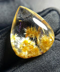 A teardrop-shaped clear resin cabochon containing golden-yellow dandelion heads, resting on a dark textured fabric background.