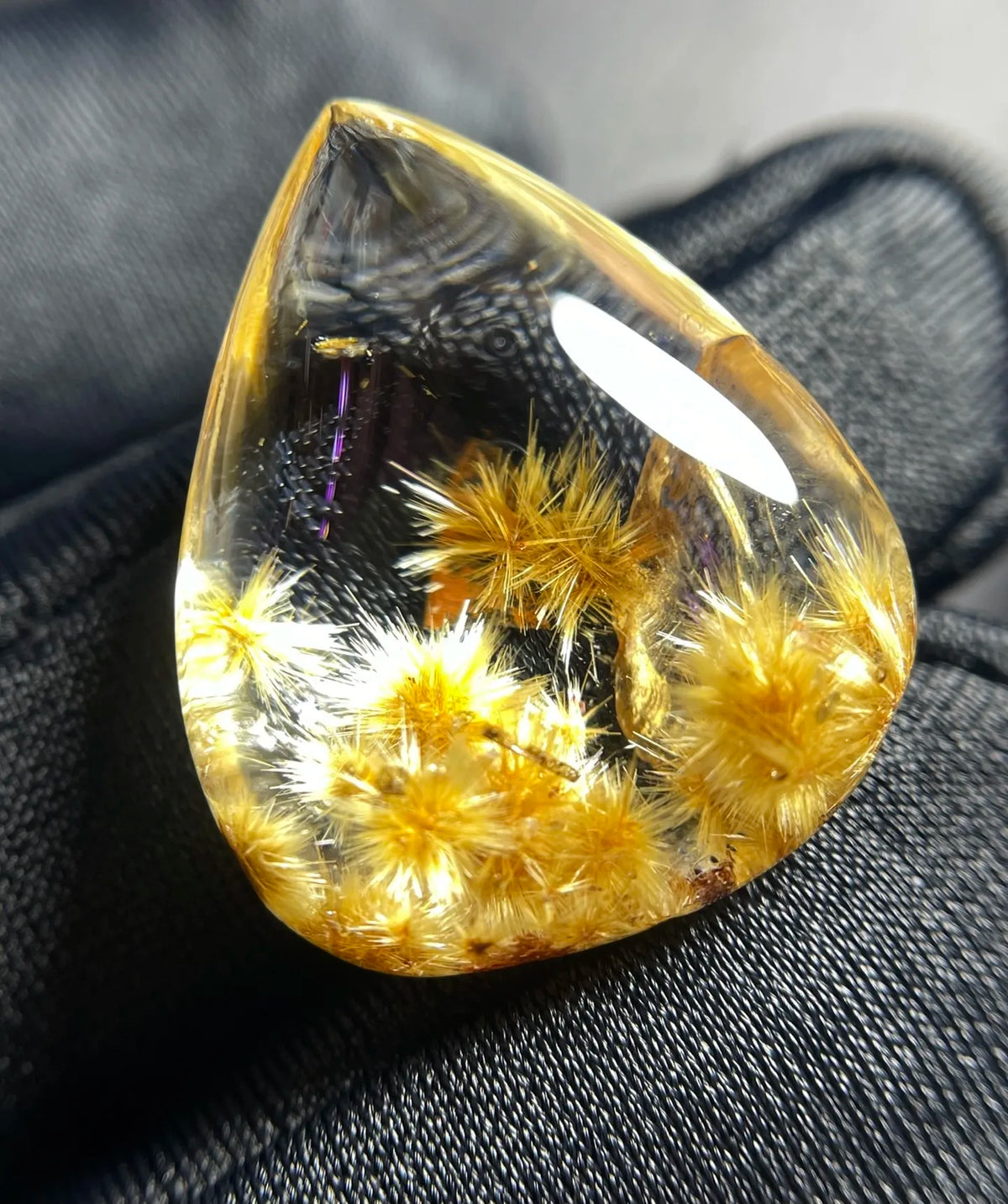 A teardrop-shaped clear resin cabochon containing golden-yellow dandelion heads, resting on a dark textured fabric background.