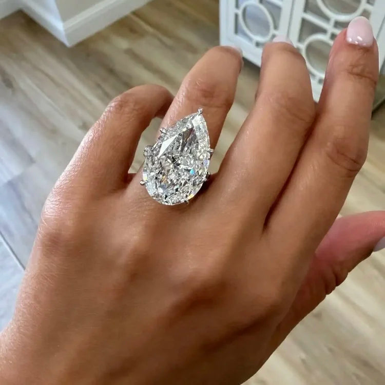 A large pear-cut diamond ring set on a silver-toned band, worn on a person's hand with a light wood floor and white furniture in the background.
