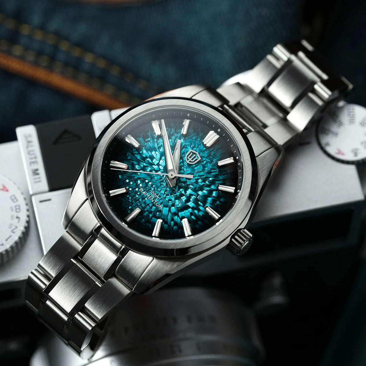 A stainless steel wristwatch with a textured turquoise and black floral dial, resting on a camera body with a denim fabric background.