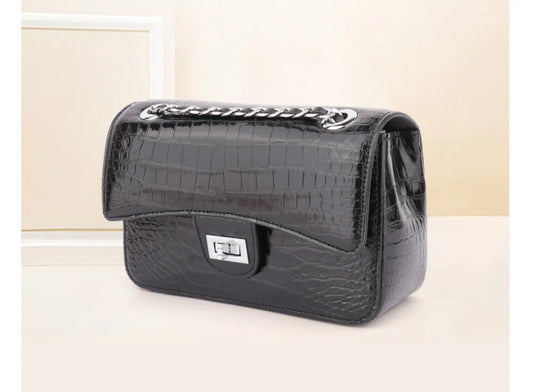 A black crocodile-embossed leather handbag with a silver-tone chain strap and a silver turn-lock clasp, displayed on a light beige surface.