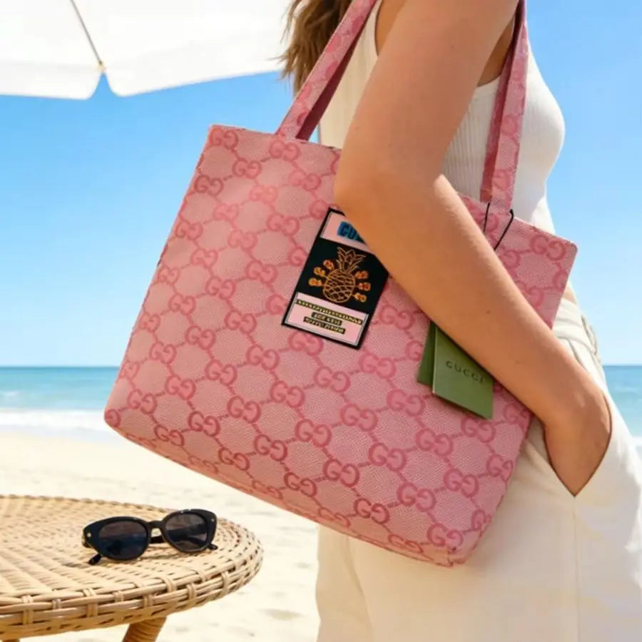 A pink Gucci tote bag with the brand's signature GG monogram pattern, carried by a woman on a beach. The bag features a black and gold pineapple patch and a green Gucci tag, set against a background of blue sky and ocean.