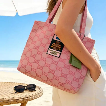 A pink Gucci tote bag with the brand's signature GG monogram pattern, carried by a woman on a beach. The bag features a black and gold pineapple patch and a green Gucci tag, set against a background of blue sky and ocean.