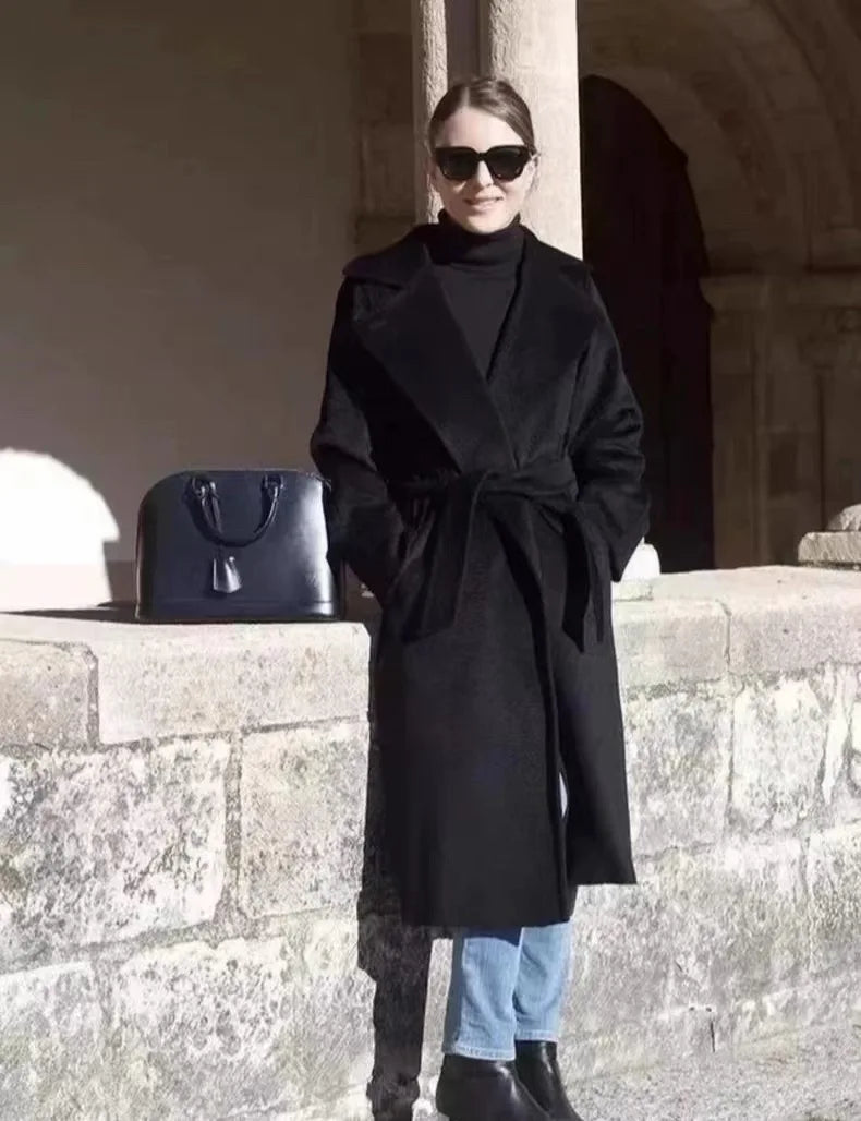 A woman wearing a long black coat over a black turtleneck, light blue jeans, and black boots, standing outdoors next to a black handbag against a stone wall and archway.