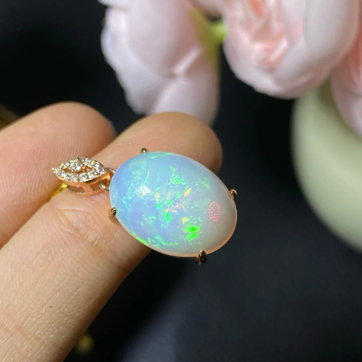 A close-up of a hand holding a rose gold ring with a large oval cabochon opal featuring iridescent blue, green, and pink flashes, set against a dark background with soft pink flowers.