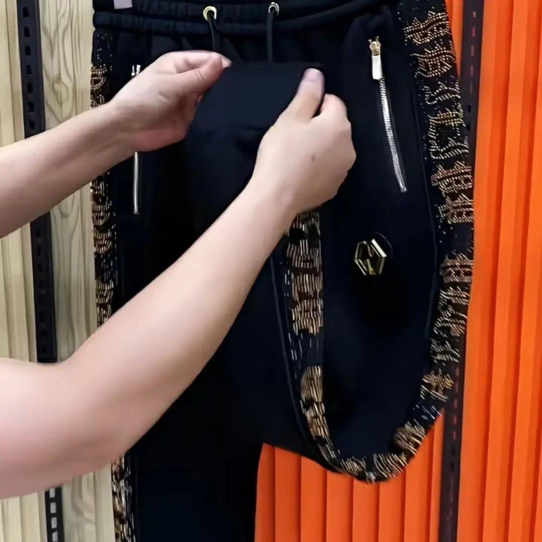 Black pants with gold patterned side panels, gold zippers, and a gold geometric metal accent, shown being held by a person's hands against an orange and wood background.