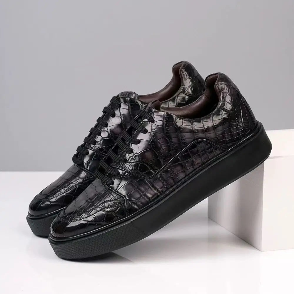 A pair of black low-top sneakers with a glossy crocodile-embossed texture, black laces, and chunky black platform soles, displayed on a white block against a gray background.