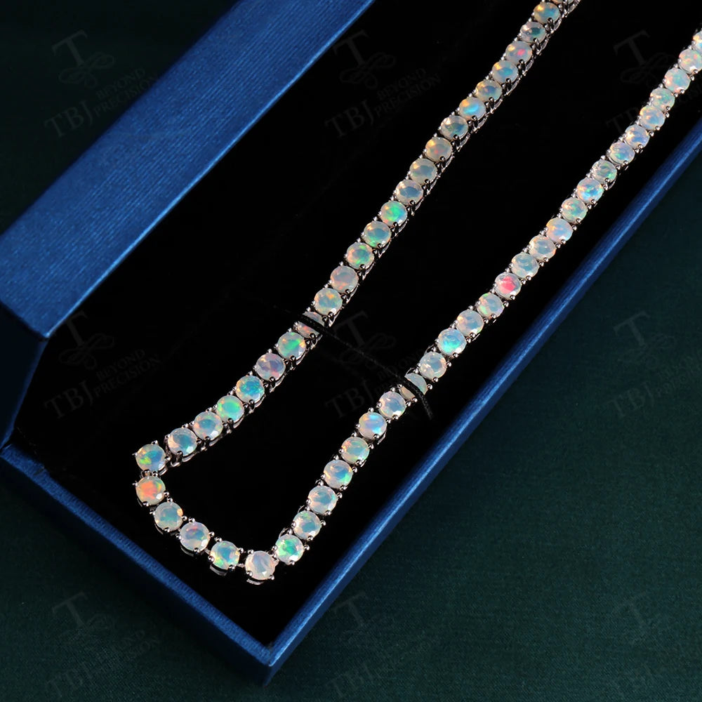 A silver-tone tennis necklace set with round iridescent opal gemstones, displayed inside an open dark blue presentation box on a dark green background.