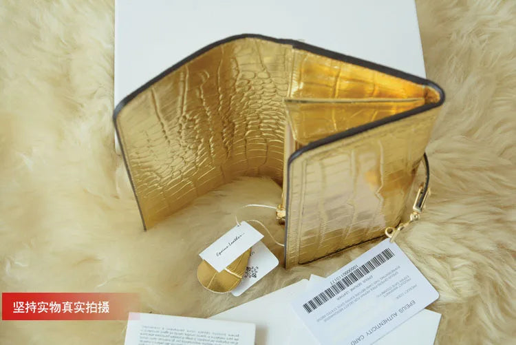 A gold crocodile-embossed leather wallet or clutch shown open on a white surface, with a white faux-fur surface underneath and product tags nearby. The item has a metallic gold finish with dark trim and a small gold zipper pull.