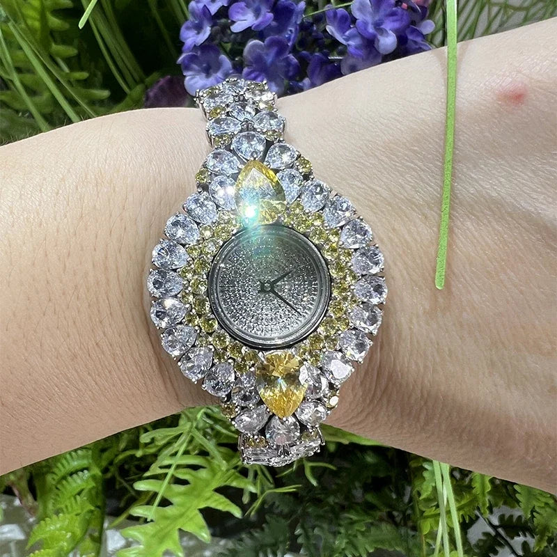 A close-up of a wrist wearing a heavily embellished watch with clear rhinestones and gold-tone accents, set against a background of green foliage and purple flowers.